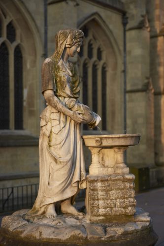 Statue of a girl in white marble pouring water from a vase into a shallow bowl outside Bath Abbey in the historic Georgian city of Bath in Somerset, England